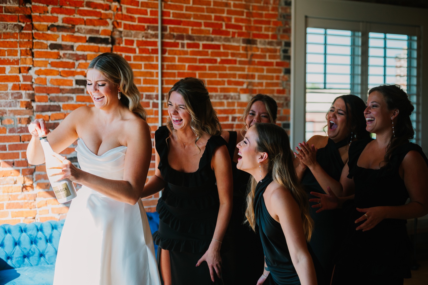 Bride opening a bottle of champagne with her bridesmaids laughing behind her