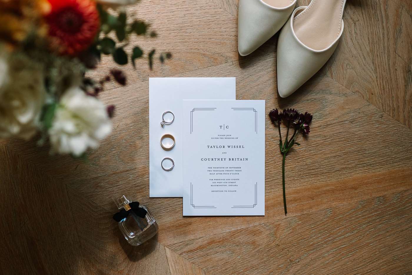 Flatlay of a modern black and white wedding invitation with flowers, perfume, and white pointed-toe bridal shoes